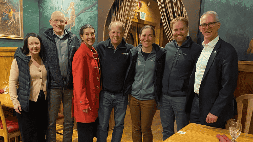 Bob Utah Indian 122025 Group photo taken in a restaurant featuring seven people standing side by side. From left to right: a woman in a beige sweater and vest, a man in a blue shirt and jacket, a woman in a red coat, a man in a black jacket, a woman in a gray jacket, a man in a gray jacket, and Bob Inglis wearing a suit and glasses. They are smiling and standing in front of a decorative curtain and painted wall.
