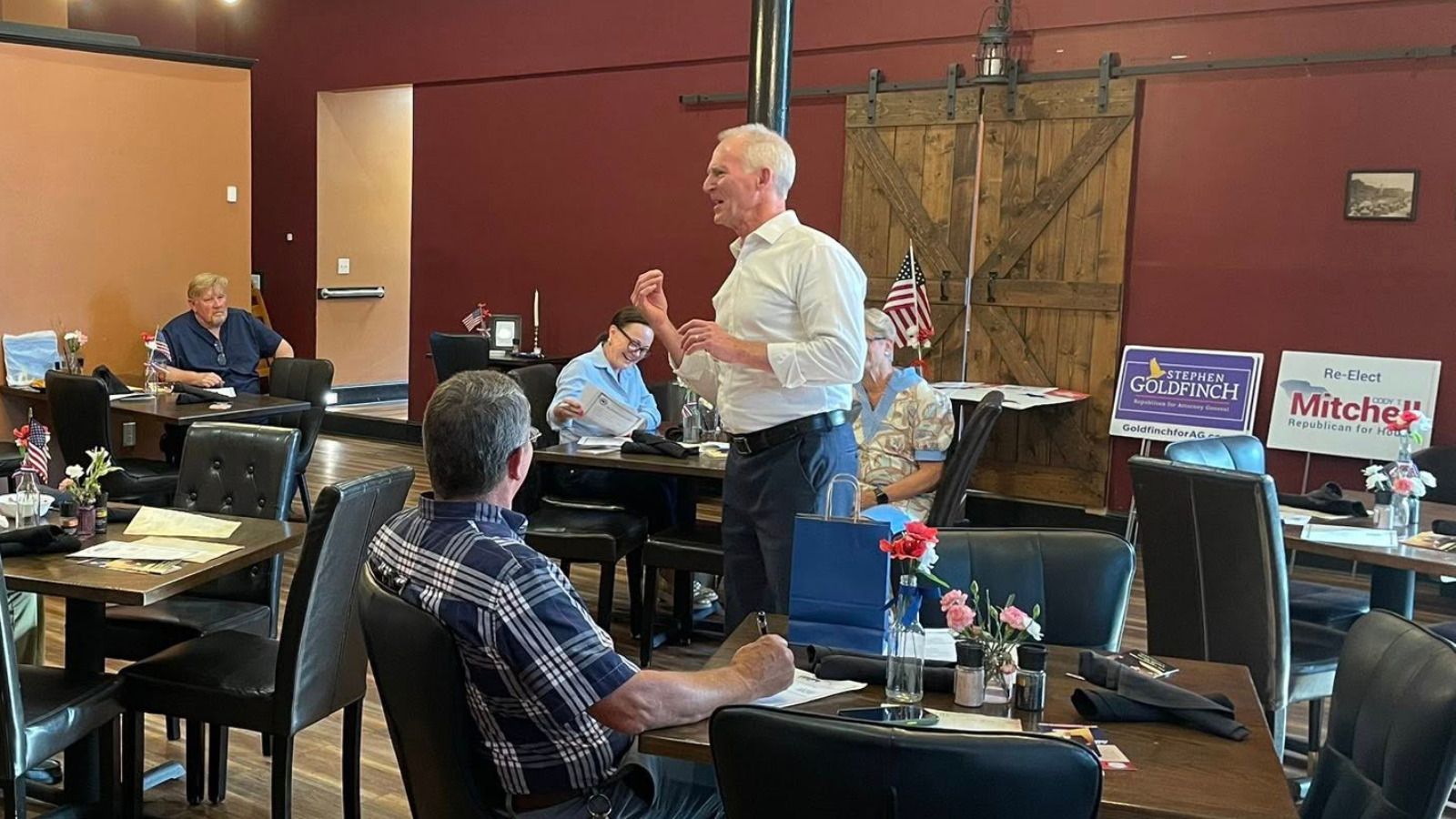 Darlington Co GOP Bob Inglis 2026 Bob Inglis stands and speaks to members of the Darlington County Republican Party gathered in a restaurant meeting room in Hartsville, South Carolina. Attendees sit at tables listening and taking notes as Inglis discusses conservative approaches to climate and energy policy. Campaign signs and American flags are visible in the background.