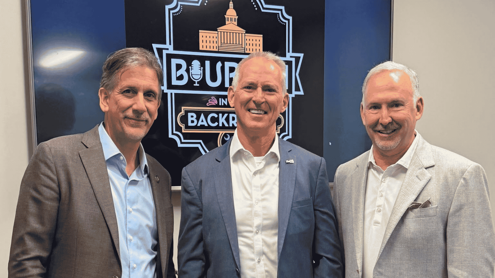 Bob Inglis Joins Bourbon in the Back Room Podcast Former Congressman Bob Inglis stands between podcast hosts Vincent Sheheen and Joel Lourie in front of a Bourbon in the Back Room sign. All three are smiling and dressed in business attire before recording an episode discussing conservative approaches to addressing climate change, free-market solutions, and carbon pricing.